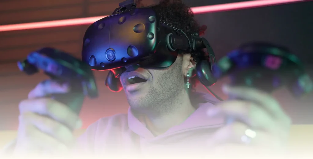 Egy virtuális valóság-sisakot viselő személy két vezérlőt tart a kezében, és láthatóan elragadtatva és meglepődve figyel. Lila kapucnis pulóvert visel, a háttérben színes fények láthatók